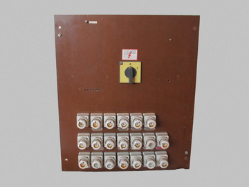 Front view of the fuse box. It's a large rectangular board made of a smooth light brown material. To it are mounted 20 fuse holders made of white ceramic and a power switch on the top. On the left and right of the switch there are signs of two pieces of equipment that were once installed.