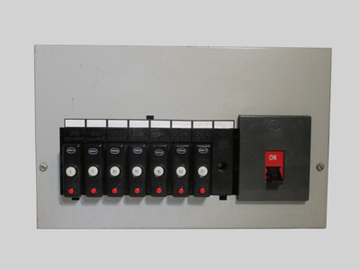Front view of the MEM breaker box. It's made of light grey metal. Eight circuit spaces are present, of which seven are occupied by breakers, made of black plastic. To the right, there is the main switch, with a red label indicating if it's on or off.