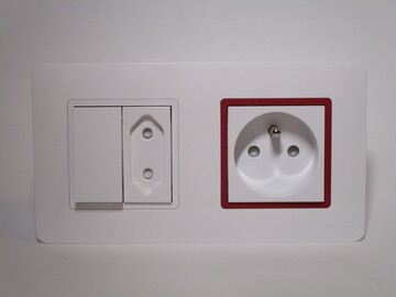 Front view of a double-wide electrical fitting, with a lightswitch and non-earthed socket on the left and an earthed socket on the right. The earthed socket has a red surround on the cover frame which is otherwise white.